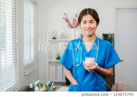 Cheerful nurse in blue scrubs with stethoscope holding a piggy bank, symbolizing healthcare savings in a bright medical office. Cheerful nurse in blue scrubs with stethoscope holding a piggy bank, symbolizing healthcare savings in a bright medical office. 124071705
