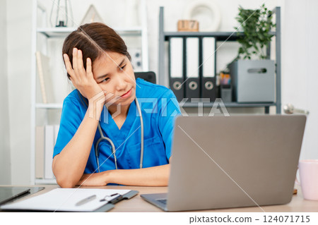 Exhausted healthcare worker in blue scrubs using a laptop at a desk, surrounded by medical documents and office supplies. 124071715