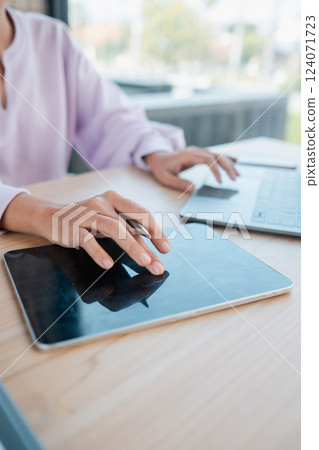A person interacts with a digital tablet and laptop on a wooden desk, showcasing modern technology in a bright workspace. 124071723