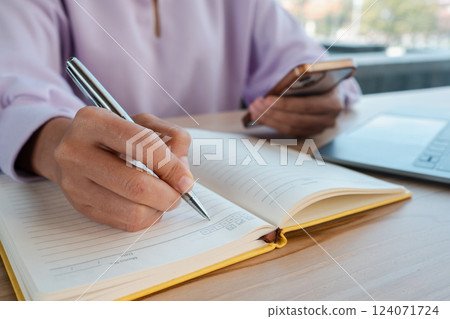Close-up of a person writing in a notebook while holding a smartphone, showcasing multitasking in a modern workspace. 124071724
