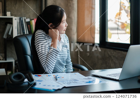 Young woman in a striped shirt sits at a desk with a laptop and documents, looking thoughtfully out the window. 124071736