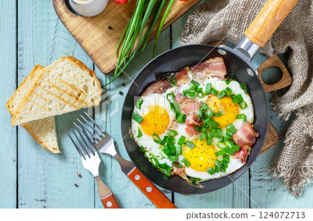 Spring scrambled eggs with green onions and bacon in a cast iron skillet on a rustic wooden background. Top view flat lay. Spring scrambled eggs with green onions and bacon in a cast iron skillet on a rustic wooden background. Top view flat lay. 124072713
