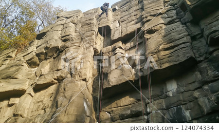 Rock climber descends a vertical cliff face during a clear day in an outdoor climbing area surrounded by trees 124074334
