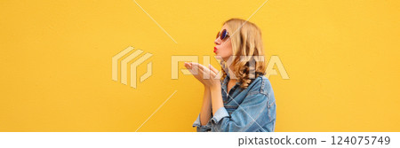 Beautiful stylish young woman blowing kiss in glasses, denim jacket posing on yellow wall background 124075749