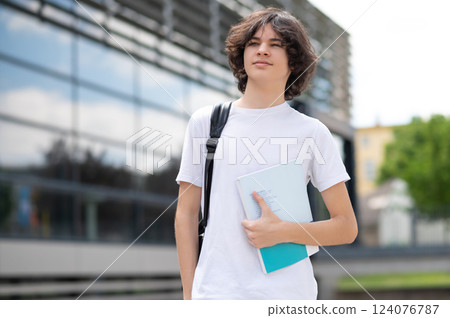 Teenager in casual outfit walking on city street carrying study bag and book 124076787