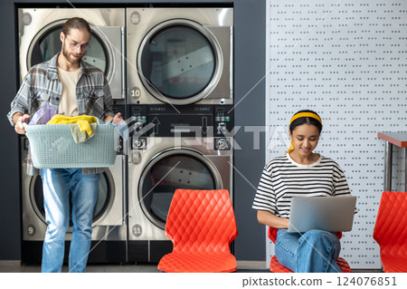 Woman in laundromat using laptop while man using washing machine for washing clothing 124076851