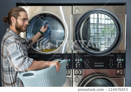 Man in laundromat using commercial washing machine while completing his laundry chores Man in laundromat using commercial washing machine while completing his laundry chores 124076853