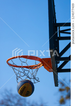 Basketball in Motion: A Close-Up View of a Wilson Basketball About to Score Through an Orange Hoop Under a Clear Blue Sky 124077489