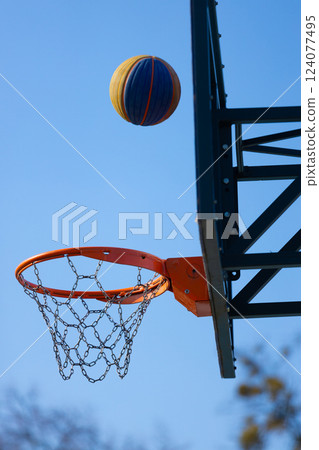 A Vibrant Basketball Soars Towards the Hoop in Bright Blue Sky, Illustrating the Thrill of Outdoor Sports and the Joy of Playing Basketball with Friends and Family Gathered Nearby A Vibrant Basketball Soars Towards the Hoop in Bright Blue Sky, Illustrating the Thrill of Outdoor Sports and the Joy of Playing Basketball with Friends and Family Gathered Nearby 124077495