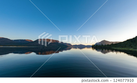As the sun sets behind the distant mountains, its golden light casts a warm glow on the rugged cliffs, beautifully reflected on the lake's calm surface. The sky transitions from deep blue to soft As the sun sets behind the distant mountains, its golden light casts a warm glow on the rugged cliffs, beautifully reflected on the lake's calm surface. The sky transitions from deep blue to soft 124077910
