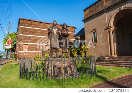 A bronze statue of a man and woman stands on a stone pedestal near a historic building with arched entrances, surrounded by tropical plants and a lawn. 124078234