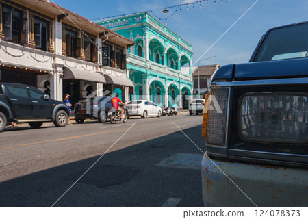 Turquoise colonial style building with arched windows in Phuket, Thailand, surrounded by parked vehicles, a scooter rider, and string lights overhead. 124078373