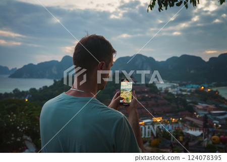 A man photographs a tropical landscape with limestone cliffs, rooftops, and a cloudy sky in Phuket, Thailand. Soft light highlights the karst formations. 124078395