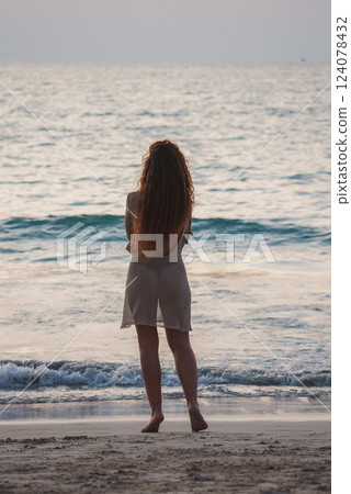 A barefoot woman with long red hair stands on a sandy beach, gazing at the calm ocean. Gentle waves and soft lighting create a tranquil scene. 124078432