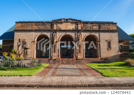 The Kauai Museum in Lihue, Hawaii features a classical facade with three arches, ornate columns, a statue, brick pathways, and a clear blue sky. 124078441