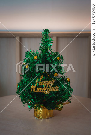 Small Christmas tree adorned with golden ornaments and a glittery Happy New Year sign, set on a light wooden table with a warm, festive background. Small Christmas tree adorned with golden ornaments and a glittery Happy New Year sign, set on a light wooden table with a warm, festive background. 124078490