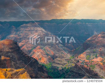 Waimea Canyon on Kauai Island features rugged cliffs with red, orange, and green layers, illuminated by golden sunlight under a partly cloudy sky. 124078504