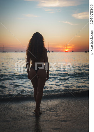 A woman with curly hair faces a vibrant sunset over calm ocean waves, with silhouetted sailboats on the horizon in Phuket or Koh Phi Phi islands. 124078506