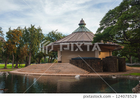A circular pavilion with a conical roof, stone steps, and columns, surrounded by lush greenery, tall trees, and a serene pond in a Hawaii park. 124078529