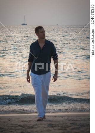 A man in a dark shirt and white pants walks along a tropical beach at sunset. Gentle waves and a sailboat on the calm sea create a serene scene. 124078556