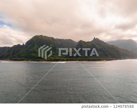 Lush green cliffs of the Napali Coast rise above a sandy beach with gentle waves, under a mix of cloudy and sunny skies on Kauai Island, Hawaii. Lush green cliffs of the Napali Coast rise above a sandy beach with gentle waves, under a mix of cloudy and sunny skies on Kauai Island, Hawaii. 124078588
