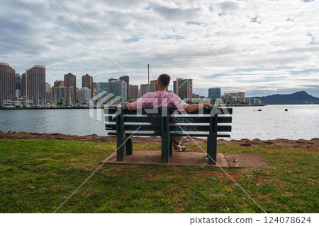 A man sits on a green bench by a grassy area, facing the Honolulu skyline, a marina with docked boats, and Diamond Head under a partly cloudy sky. 124078624