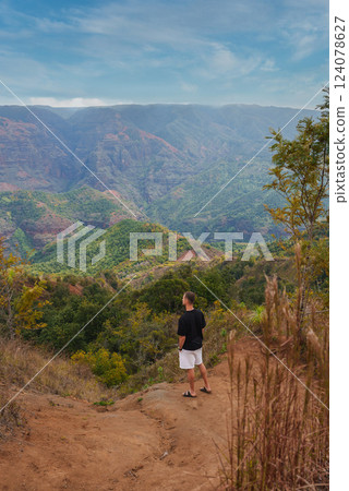 A man stands on a dirt trail with Waimea Canyon's rugged cliffs, red and green layers, and scattered trees under a clear blue sky in Kauai, Hawaii. A man stands on a dirt trail with Waimea Canyon's rugged cliffs, red and green layers, and scattered trees under a clear blue sky in Kauai, Hawaii. 124078627