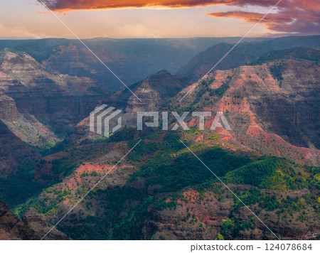 Aerial view of Waimea Canyon on Kauai Island, Hawaii, featuring multicolored cliffs, lush vegetation, and a dramatic orange and purple sky. 124078684