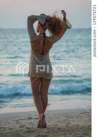 A woman in a light dress stands barefoot on a beach, facing the ocean at sunset or sunrise. Calm waves meet the shore in a serene tropical setting. 124078721