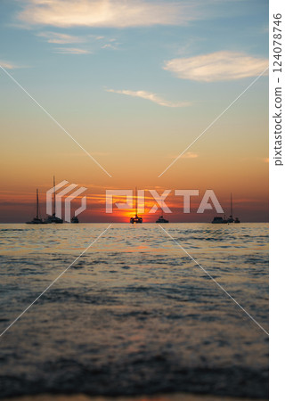 Sunset over the ocean near Phuket or Koh Phi Phi islands, Thailand, with sailboats silhouetted against orange and pink hues and gentle sea ripples. 124078746