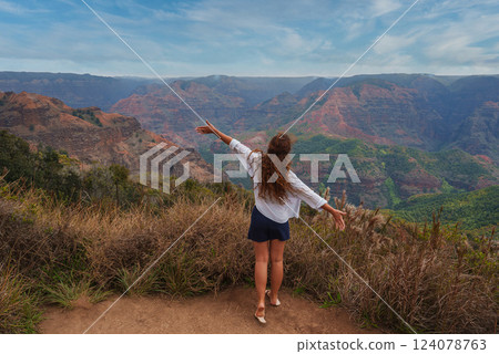 A woman stands on a dirt path with arms outstretched, overlooking Waimea Canyon's red and green cliffs, lush vegetation, and rugged terrain under a cloudy sky. 124078763