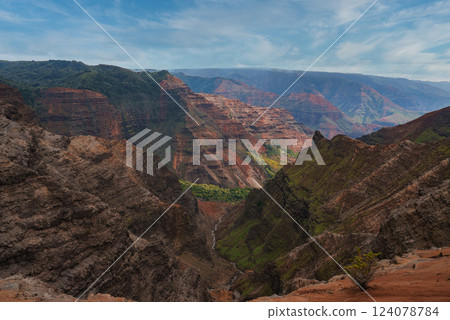 Rugged cliffs in red, green, and brown dominate Waimea Canyon on Kauai. A narrow stream flows through the canyon floor under a clear sky with clouds. Rugged cliffs in red, green, and brown dominate Waimea Canyon on Kauai. A narrow stream flows through the canyon floor under a clear sky with clouds. 124078784