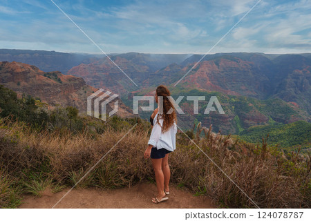 A woman with wavy hair stands on a dirt path surrounded by dry grass, gazing at Waimea Canyon's red and green cliffs under a partly cloudy sky. A woman with wavy hair stands on a dirt path surrounded by dry grass, gazing at Waimea Canyon's red and green cliffs under a partly cloudy sky. 124078787