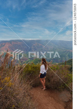A woman stands on a dirt trail surrounded by dry grass, gazing at Waimea Canyon's red and green cliffs under a partly cloudy sky on Kauai Island. 124078843