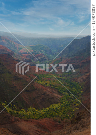 View of Waimea Canyon on Kauai, Hawaii, with red and brown cliffs, green vegetation in the valley, and a partly cloudy sky above the rugged terrain. View of Waimea Canyon on Kauai, Hawaii, with red and brown cliffs, green vegetation in the valley, and a partly cloudy sky above the rugged terrain. 124078857