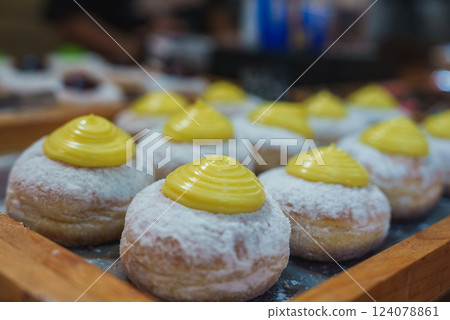 Several powdered donuts topped with bright yellow cream are arranged on a wooden tray in a bakery or market setting with a blurred background. 124078861