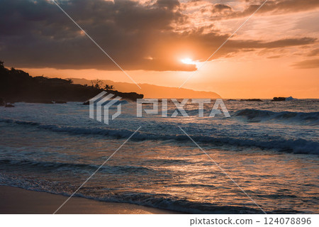 A warm orange sunset reflects on ocean waves near a Hawaiian beach, with silhouetted palm trees, rocky outcroppings, and dramatic clouds in the distance. 124078896