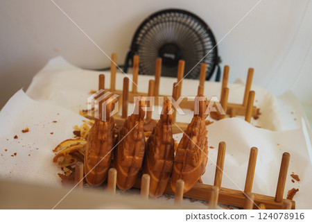 Golden brown swan shaped waffles displayed upright on a wooden rack with crumbs on white paper, a small black fan visible in the background. 124078916