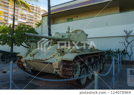 A weathered green military tank with rusted tracks is displayed outdoors near a light colored building, palm trees, and a multi story structure in Hawaii. 124078969