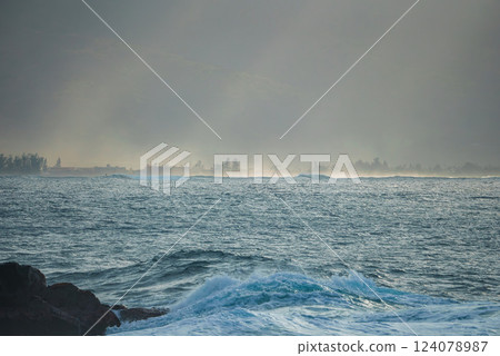 Gentle waves meet a misty shoreline with tree and building silhouettes. The soft lighting suggests early morning or late afternoon on a Hawaiian island. 124078987