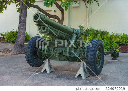 A green military artillery cannon rests on white stands outdoors, surrounded by tropical vegetation and a light colored building wall in Hawaii. 124079019