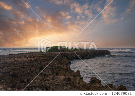 A jagged coastline on a Hawaiian island with rocky terrain, a small green patch, calm ocean waves, and a sky in warm orange and pink hues. A jagged coastline on a Hawaiian island with rocky terrain, a small green patch, calm ocean waves, and a sky in warm orange and pink hues. 124079081