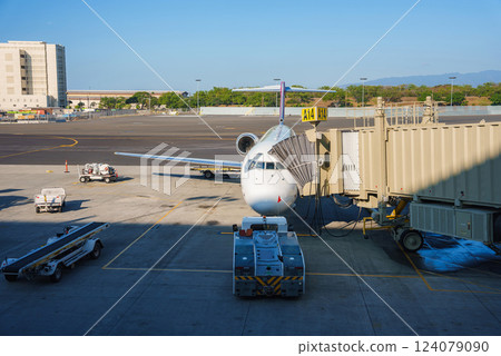 A parked airplane at gate A14 with a jet bridge, baggage carts, and ground equipment on a sunny day. Mountains and trees are visible in the distance. 124079090