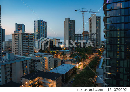 Modern high rise buildings and a construction crane in Honolulu during twilight, with warm lights, tropical trees, and the ocean faintly visible. 124079092