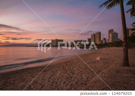 Serene view of Waikiki Beach in Honolulu during sunset, featuring a lone palm tree, a resting dog, and high rise buildings reflecting soft hues. 124079203