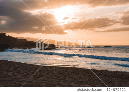 A serene Hawaiian beach at sunset with golden light filtering through clouds, rocky cliffs in the distance, and gentle waves along the sandy shore. 124079321