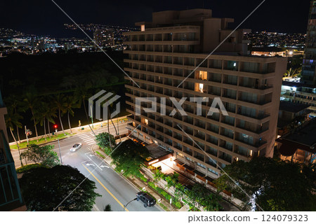 A multi story building with lit windows near a quiet street intersection, lined with palm trees. City lights and a dark water body are visible in the background. A multi story building with lit windows near a quiet street intersection, lined with palm trees. City lights and a dark water body are visible in the background. 124079323