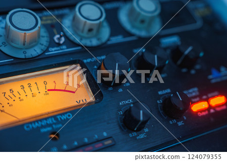 The image shows a detailed view of an audio mixing console, featuring various knobs, dials, and a glowing VU meter, indicating audio levels in a studio setting. The image shows a detailed view of an audio mixing console, featuring various knobs, dials, and a glowing VU meter, indicating audio levels in a studio setting. 124079355