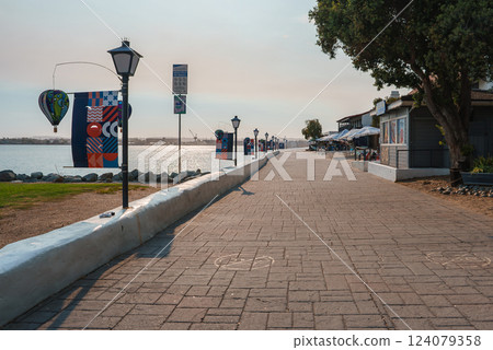A scenic waterfront promenade features street lamps and colorful banners. Small shops with outdoor seating line the path, with calm water and distant structures visible. A scenic waterfront promenade features street lamps and colorful banners. Small shops with outdoor seating line the path, with calm water and distant structures visible. 124079358