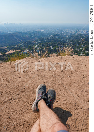 A person sits on a dirt path with legs stretched out, overlooking Los Angeles skyline from a high vantage point on a clear, slightly hazy day. 124079361